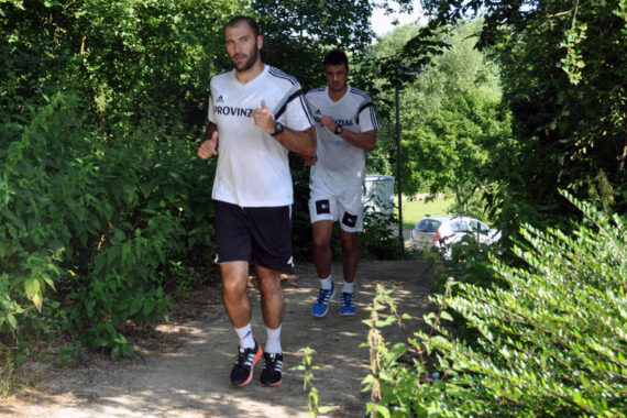 Zwei Männer in weißen Hemden und schwarzen Shorts joggen auf einem schattigen Feldweg, der von grünen Bäumen und Sträuchern umgeben ist, wobei das Sonnenlicht durch die Blätter fällt. Ein weißes Auto ist teilweise im Hintergrund zu sehen.