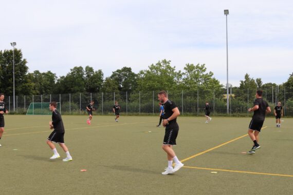 Eine Gruppe von Menschen in schwarzer Sportkleidung joggt und trainiert auf einem Sportplatz im Freien, der von einem Zaun und Bäumen umgeben ist, unter einem teilweise bewölkten Himmel.