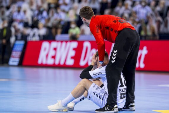 Ein Handballspieler sitzt auf dem Spielfeld und sieht verzweifelt aus, während ein Mannschaftskamerad in einem roten Trikot neben ihm steht und ihm Unterstützung anbietet. Der Hintergrund zeigt eine verschwommene Menschenmenge und eine große digitale Werbetafel.
