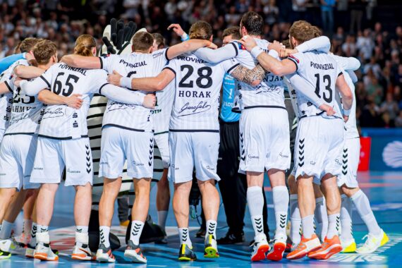 Eine Handballmannschaft in weißen Trikots steht auf einem Hallenplatz zusammen, die Arme umeinander gelegt, im Hintergrund ist eine Zuschauermenge zu sehen.