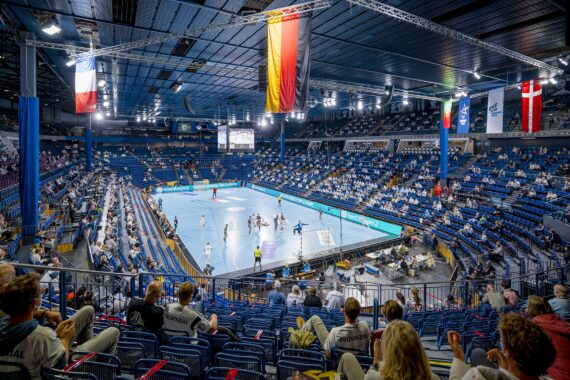 Eine große Halle mit Zuschauern, die ein Handballspiel auf einem blauen Spielfeld verfolgen. Fahnen hängen von der Decke, und die Tribünen sind teilweise mit sitzenden Fans gefüllt. Helle Lichter beleuchten die Spieler auf dem Spielfeld.