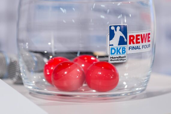 Eine durchsichtige Schale mit vier roten Bällen im Inneren, die auf der Vorderseite die Logos der DKB Handball-Bundesliga und des REWE Final Four tragen. Neben der Schale sind zwei Mikrofone zu sehen.