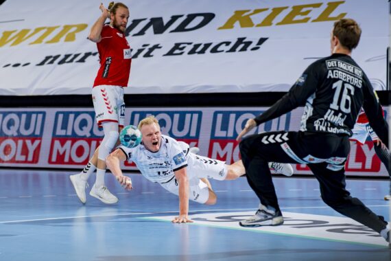 Ein Handballspieler in einem weißen Trikot springt mit einem Ball nach vorne, während ein Torwart in Schwarz sich darauf vorbereitet, den Schuss zu blocken; ein weiterer Spieler in Rot befindet sich im Hintergrund. Hinter ihnen sind ein großes Banner und Sponsorenlogos zu sehen.