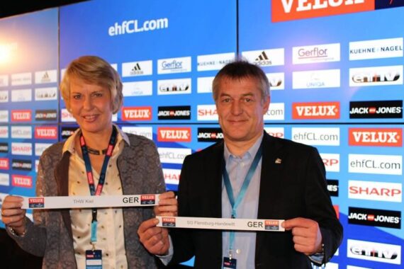 Zwei Personen stehen vor einem Sponsorenhintergrund und halten jeweils einen Papierstreifen mit den Namen deutscher Handballmannschaften: THW Kiel und SG Flensburg-Handewitt. Sie befinden sich bei einer professionellen Sportveranstaltung.