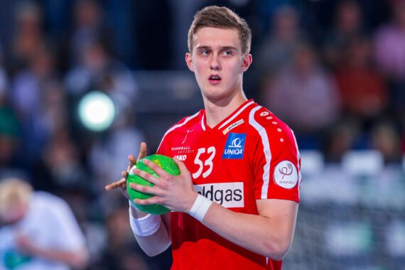Ein männlicher Handballspieler in einem roten Trikot, der einen grünen Ball in der Hand hält, blickt während eines Spiels konzentriert auf eine unscharfe Menschenmenge im Hintergrund.