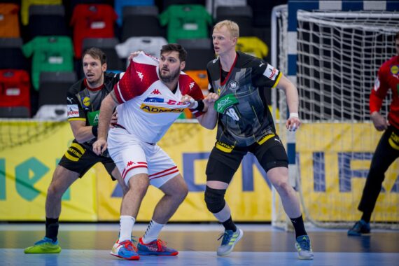 Drei männliche Handballspieler treten in der Nähe des Tores gegeneinander an. Ein Spieler in einem weiß-roten Trikot wird eng von zwei Gegnern in schwarzen Trikots bewacht, die sich alle auf das Spielgeschehen konzentrieren, wobei im Hintergrund bunte Hemden zu sehen sind.