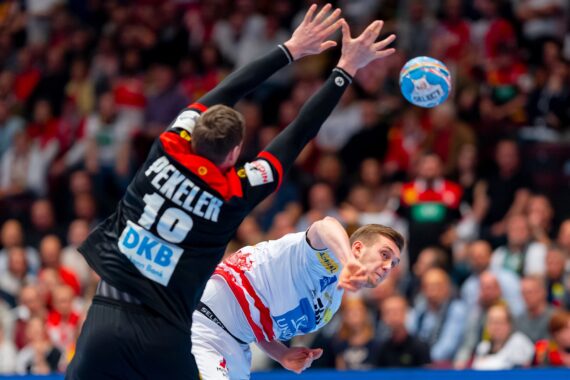 Ein Handballspieler in einem weiß-roten Trikot springt, um den Ball zu werfen, während ein Spieler in einem schwarzen Trikot mit erhobenen Armen versucht, den Ball zu blocken. Im unscharfen Hintergrund ist eine Zuschauermenge zu sehen.