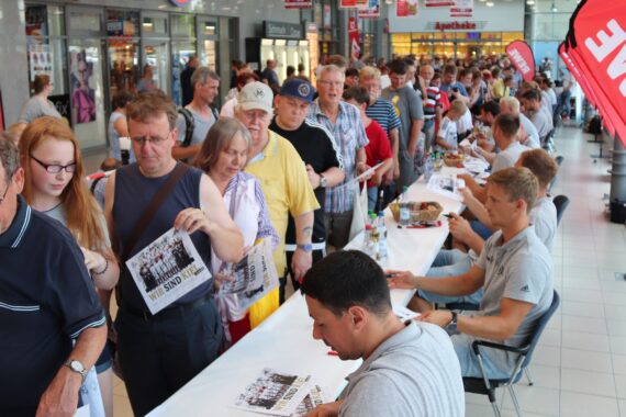 Eine lange Schlange von Menschen wartet in einem Einkaufszentrum auf ein Autogramm von vier Männern, die an Tischen sitzen, während im Hintergrund REWE-Banner und -Märkte zu sehen sind.