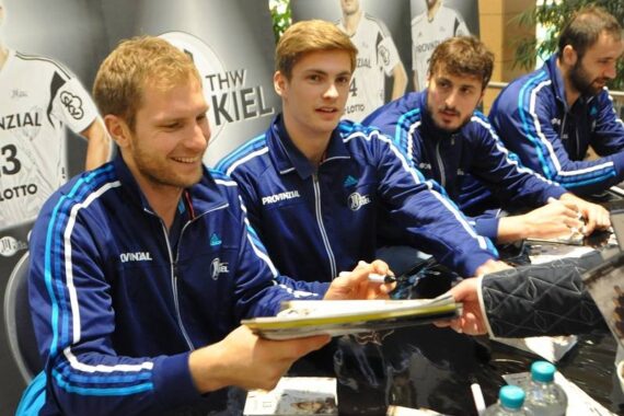 Vier männliche Sportler in passenden blauen Trainingsanzügen sitzen an einem Tisch und geben Autogramme für Fans. Ein Sportler gibt einen signierten Gegenstand zurück, während andere ebenfalls mit den Fans ins Gespräch kommen. Auf dem Tisch sind Sportposter und Wasserflaschen zu sehen.