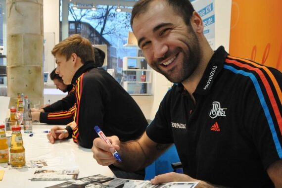 Ein lächelnder Mann in einem schwarzen Sporthemd unterschreibt an einem Tisch ein Autogramm mit einem Marker. Neben ihm sitzen zwei weitere Personen, die ebenfalls Autogramme geben. Flaschen und Fotos sind auf dem Tisch verteilt.