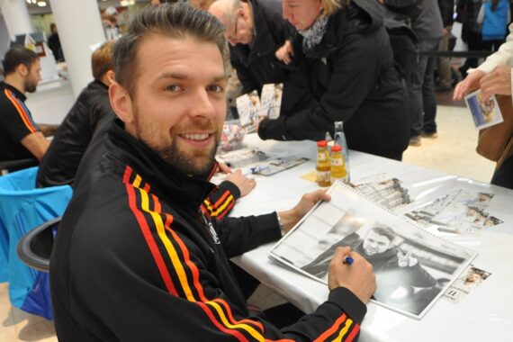 Ein Mann in einer schwarz-roten Sportjacke lächelt in die Kamera, während er an einem Tisch ein Foto signiert; mehrere Personen stehen in der Nähe und warten mit Gegenständen in der Hand.