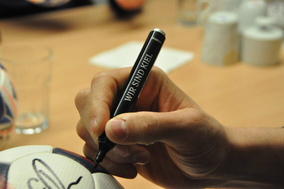 Eine Nahaufnahme einer Hand, die mit einem schwarzen Marker mit der Aufschrift WIR SIND KIEL einen weißen Fußball signiert. Der Hintergrund zeigt einen Tisch mit Tassen, Papier und einem unscharfen Glas.