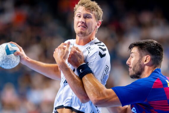 Ein Handballspieler in einem weißen Trikot hält den Ball, während er von einem Gegner in einem blau-roten Trikot während eines intensiven Spiels eng verteidigt wird.