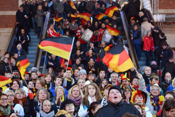 Eine große Menschenmenge, von denen viele deutsche Fahnen schwenken und Kleidung und Accessoires mit deutschen Motiven tragen, versammelt sich vor einer Rolltreppe und wirkt aufgeregt und feierlich.