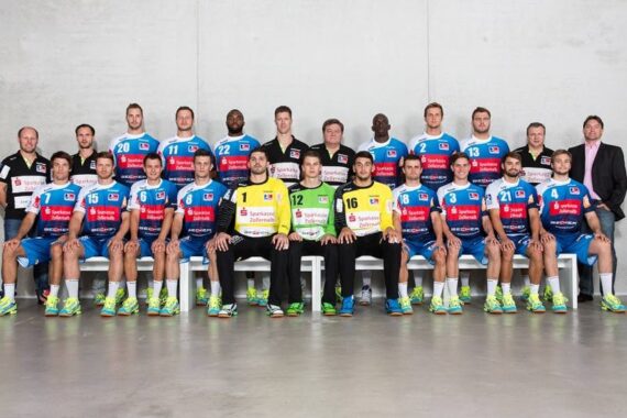 Eine Herren-Handballmannschaft stellt sich in einer Halle für ein formelles Gruppenfoto auf. Die Spieler tragen blaue Trikots mit roten und weißen Akzenten, während die drei in der Mitte gelbe oder grüne Torwarttrikots tragen. Trainer und Betreuer stehen hinter ihnen vor einem schlichten grauen Hintergrund.