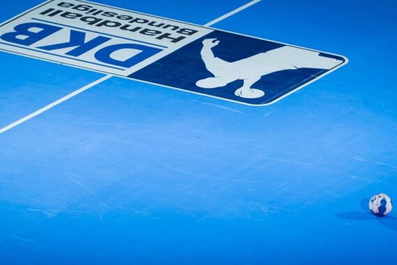 Ein einzelner Handball liegt auf einem leuchtend blauen Hallenfeld, neben einem großen, auf den Boden gemalten DBK Handball-Bundesliga-Logo. Der Raum ist offen und minimalistisch.