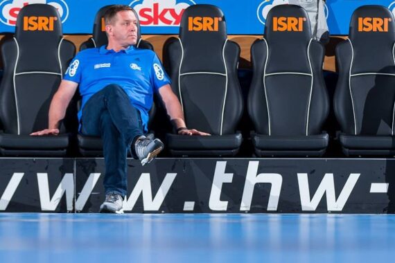 Ein Mann in blauem Hemd und Jeans sitzt allein auf einer Reihe schwarzer Sportstühle mit der Aufschrift ISRI auf den Kopfstützen. Der Hintergrund zeigt das sky-Logo und einen blauen Boden davor.