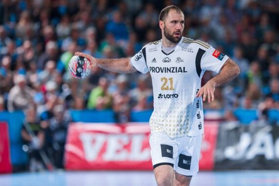 Ein männlicher Handballspieler in einem weißen Trikot mit der Nummer 21 hält den Ball während eines Spiels in der rechten Hand. Der Hintergrund zeigt eine unscharfe Menge und Banner.