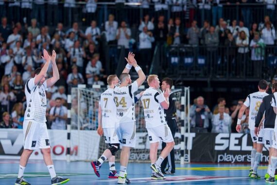 Handballspieler in weißen Trikots klatschen und winken dem Publikum nach einem Spiel in einer Halle zu, im Hintergrund sind Zuschauer zu sehen.