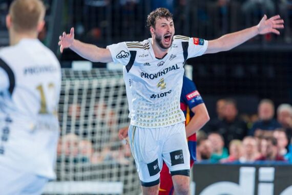 Ein männlicher Handballspieler in einem weißen Trikot jubelt mit ausgestreckten Armen vor einem Tor, während ein anderer Spieler in einem ähnlichen Trikot zu sehen ist und eine Menschenmenge im Hintergrund zusieht.