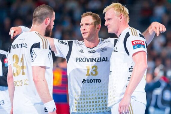 Drei männliche Handballspieler in weißen Trikots mit goldenen Details stehen zusammen auf dem Spielfeld, wobei der Spieler in der Mitte die Arme hebt und während eines Spiels zu seinen Mitspielern spricht.