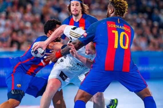 Zwei Handballspieler in blauen und roten Trikots verteidigen sich gegen einen Spieler in einem weißen Trikot, der den Ball hält, während eines intensiven Spiels. Die Zuschauer sind im Hintergrund verschwommen.