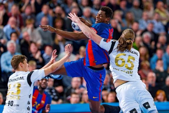 Ein Handballspieler in einem blau-roten Trikot springt, um den Ball zu schießen, während zwei Verteidiger in weißen Trikots mit den Nummern 65 und 33 versuchen, ihn während eines Spiels in einer überfüllten Arena zu blockieren.