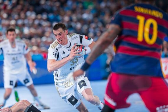 Ein Handballspieler in einem weißen Trikot mit der Nummer 33 stürmt mit dem Ball nach vorne, verfolgt von einem Gegner in einem rot-blau gestreiften Trikot mit der Nummer 10. Die Aktion findet auf einem Hallenplatz statt, mit einer unscharfen Zuschauermenge im Hintergrund.