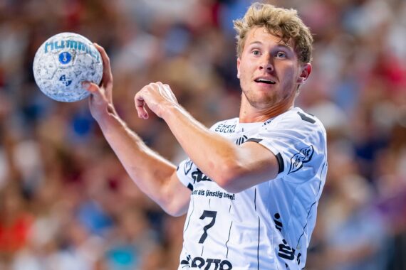Ein männlicher Handballspieler in einem weißen Trikot mit der Nummer 7 bereitet sich während eines Spiels auf den Wurf vor, während im Hintergrund eine unscharfe Menschenmenge zu sehen ist.