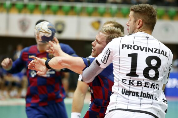 Ein Handballspieler in einem blau-roten Trikot wird während eines intensiven Spiels von einem Spieler in einem weißen Trikot mit der 18 EKBERG auf dem Rücken geblockt. Ein anderer Spieler hält den Ball im Hintergrund.