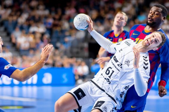Ein männlicher Handballspieler in einem weißen Trikot fällt rückwärts, während er den Ball hält, der von zwei Spielern in blauen und roten Trikots während eines energiegeladenen Spiels in einer Halle eng verteidigt wird.