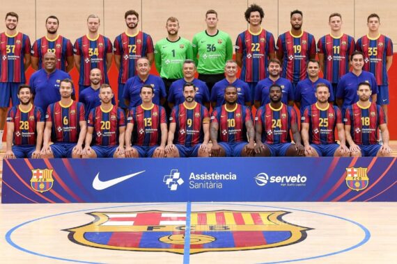 Eine Handballmannschaft der Männer in blau-rot gestreiften Trikots posiert in einer Halle für ein Gruppenfoto mit Trainern und Betreuern, die in Reihen hinter einem Schild mit den Logos der Sponsoren und einem großen Wappen des FC Barcelona auf dem Boden stehen und sitzen.