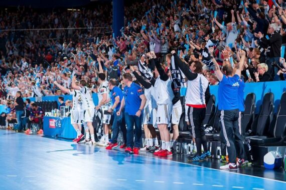 Eine jubelnde Menge jubelt und hebt die Arme in einer vollen Sporthalle, während eine Mannschaft in weiß-schwarzen Trikots während eines Handballspiels am Spielfeldrand jubelt.