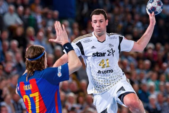 Ein Handballspieler in einem weißen Trikot springt in der Luft und bereitet sich auf einen Schuss vor, während ein Verteidiger in einem blau-roten Trikot die Hand hebt, um ihn während eines überfüllten Hallenspiels abzuwehren.