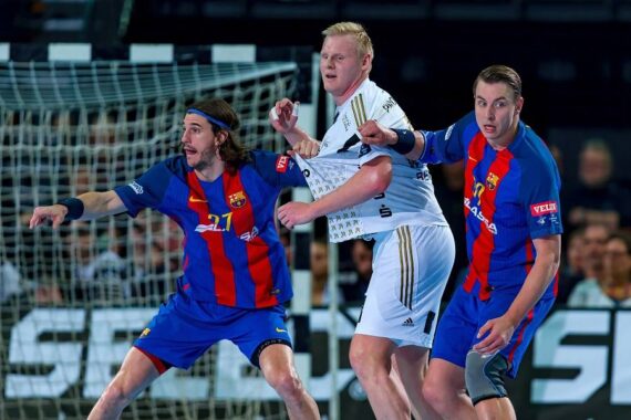 Drei männliche Handballspieler, zwei in blauen und roten Trikots und einer in einem weißen Trikot, treten während eines Spiels eng gegeneinander an, wobei der Spieler in Weiß von den beiden anderen vor einem Tor verteidigt wird.