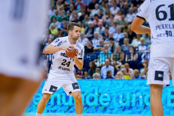 Ein männlicher Handballspieler in einem weißen Trikot hält den Ball und bereitet sich auf einen Spielzug vor, während Mannschaftskameraden und Zuschauer den Hintergrund in einer Sporthalle füllen.