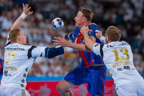 Drei männliche Handballspieler in Aktion während eines Spiels. Ein Spieler in einem blau-roten Trikot hält den Ball, während er von zwei Spielern in weißen Trikots geblockt wird, von denen einer die Nummer 13 trägt, alle mit intensiven Gesichtsausdrücken. Im Hintergrund ist ein unscharfes Publikum zu sehen.