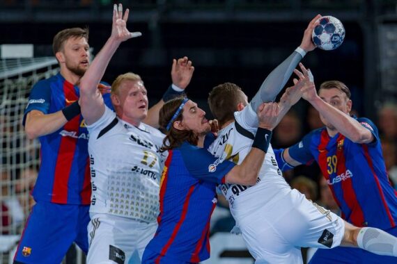 Eine Gruppe von männlichen Handballspielern zweier Mannschaften in blau-rot-weißen Trikots kämpft mit erhobenen Armen um den Ball, wobei die Spieler den Ballbesitz körperlich ausfechten.