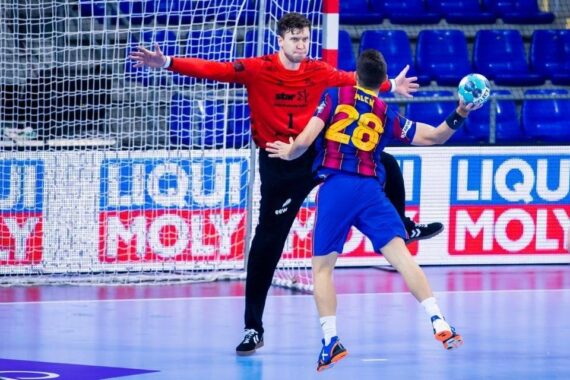 Ein Handballspieler in einem blau-roten Trikot bereitet sich auf einen Schuss vor, während ein Torwart in Rot mit weit ausgebreiteten Armen das Tor blockiert. Beide befinden sich auf einem Hallenplatz mit Werbetafeln im Hintergrund.