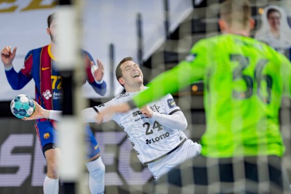 Ein Handballspieler im weißen Trikot mit der Nummer 24 versucht einen Schuss, der von einem Torhüter in einem hellgrünen Trikot geblockt wird; im Hintergrund ist ein weiterer Spieler in einem blau-roten Trikot zu sehen.
