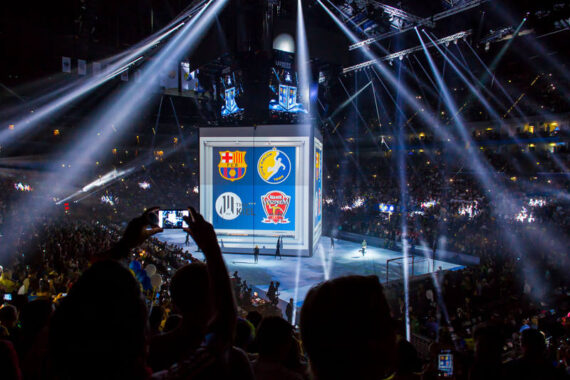 Ein großer Würfel zeigt Logos von Sportmannschaften in einer überfüllten Halle mit dramatischer Beleuchtung und Scheinwerfern, während die Zuschauer Fotos machen und das Ereignis beobachten.