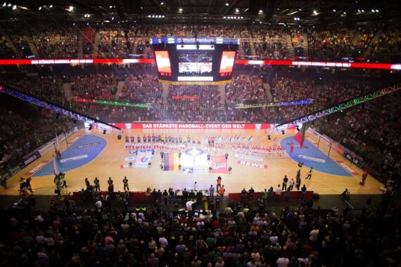 Eine überfüllte Halle mit einem hell erleuchteten Handballfeld, Mannschaften, die mit dem Gesicht zur Menge aufgereiht sind, und Fahnen, die gezeigt werden, während die Zuschauer von der Tribüne aus unter bunten Lichtern zusehen. Über dem Spielfeld ist ein Text in deutscher Sprache zu sehen.