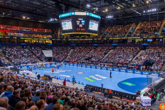 Eine überfüllte Halle mit jubelnden Zuschauern, die ein professionelles Handballspiel auf einem blauen Spielfeld verfolgen, mit einer großen Anzeigetafel über dem Spielfeld und digitalen Bannern rund um den Sitzbereich.