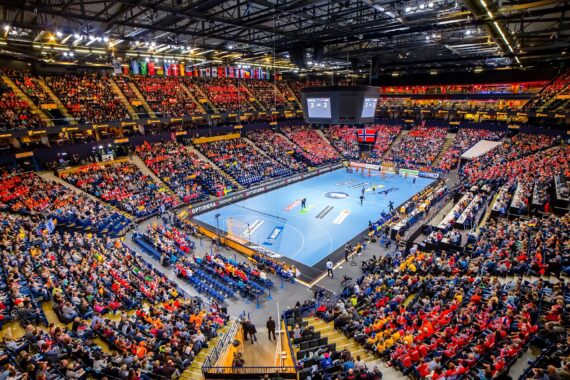 Eine überfüllte Halle mit Zuschauern, die ein Handballspiel auf einem blauen Spielfeld verfolgen, über dem die Flaggen verschiedener Länder hängen und helle Lichter die Szene erhellen.