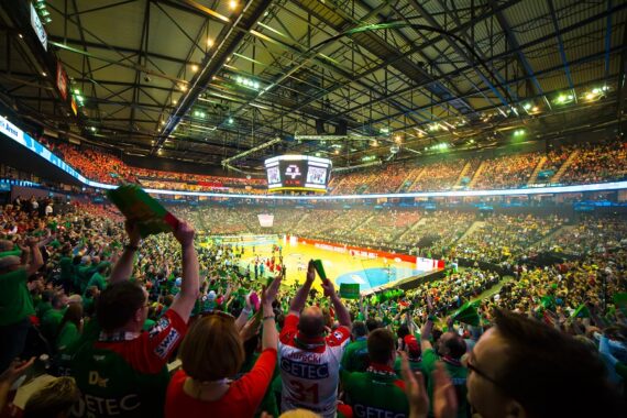 Eine große Sporthalle voller jubelnder Fans, viele in grünen Hemden, verfolgt ein intensives Spiel auf einem gut beleuchteten Platz. Das Stadion ist lebhaft, und über der Mitte hängt eine Anzeigetafel.