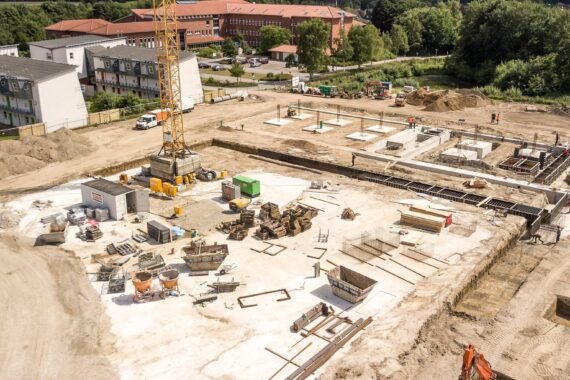 Luftaufnahme einer Baustelle mit Gebäudefundamenten, Baumaterialien, Maschinen und einem Kran. In der Nähe befinden sich Wohngebäude, grüne Bäume und ein teilweise geräumtes Gelände.