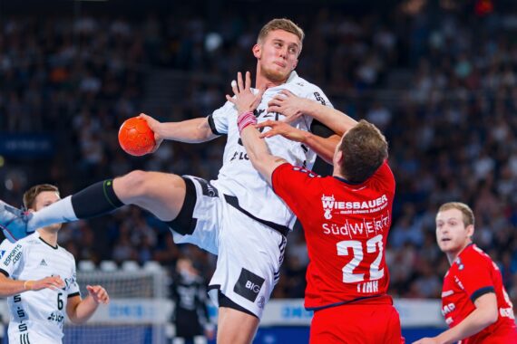 Ein Handballspieler in einem weißen Trikot springt und bereitet sich auf den Wurf vor, während ein Verteidiger in einem roten Trikot versucht, ihn während eines intensiven Spiels in einer überfüllten Halle zu blockieren.