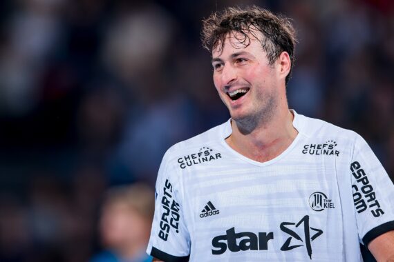 Ein männlicher Handballspieler in einem weißen Trikot lächelt während eines Spiels. Auf seinem Trikot sind verschiedene Sponsorenlogos zu sehen, darunter Star und ESPORTS.COM. Der Hintergrund ist unscharf und deutet auf eine Sporthalle hin.