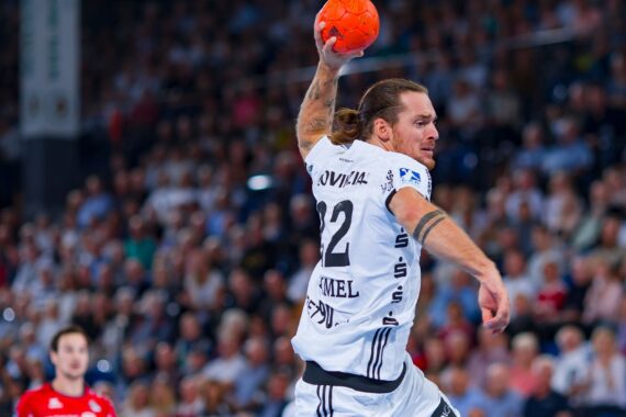 Ein männlicher Handballspieler in einem weißen Trikot mit der Nummer 22 springt in die Luft, hebt den Arm, um einen orangefarbenen Ball zu werfen, während eines intensiven Spiels mit einer unscharfen Menschenmenge im Hintergrund.