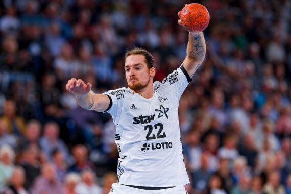 Ein männlicher Handballspieler in einem weißen Trikot mit der Nummer 22 bereitet sich darauf vor, während eines Spiels einen orangefarbenen Ball zu werfen, während im Hintergrund eine unscharfe Zuschauermenge zusieht.